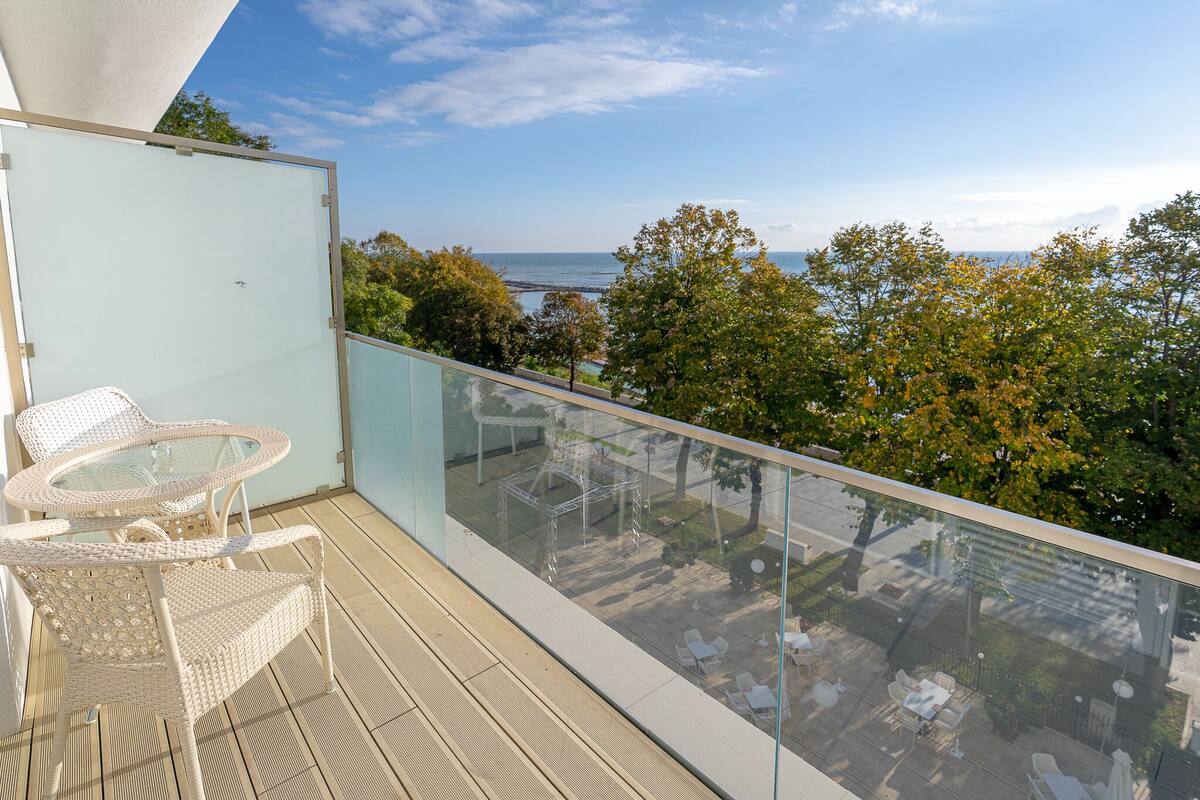 A balcony with glass railings, wicker furniture, and a view of the sea and trees.