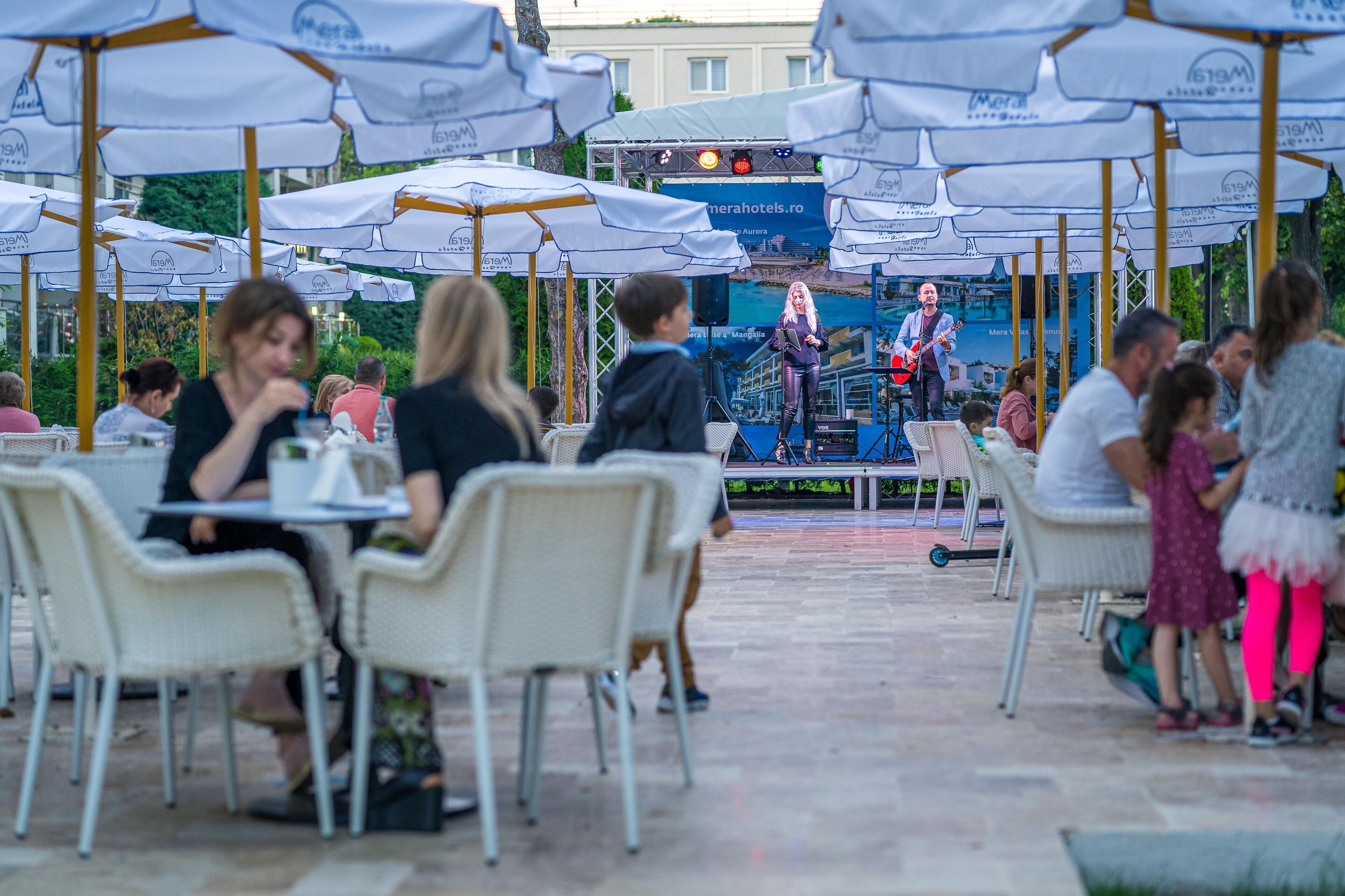 Un área de comedor al aire libre con sombrillas blancas y mesas, un escenario con un artista y un fondo con la marca del hotel.