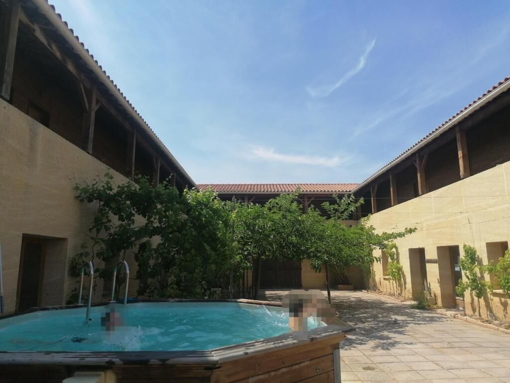 Una piscina al aire libre con solárium de madera, rodeada de edificios con tejado de tejas y un cielo despejado.