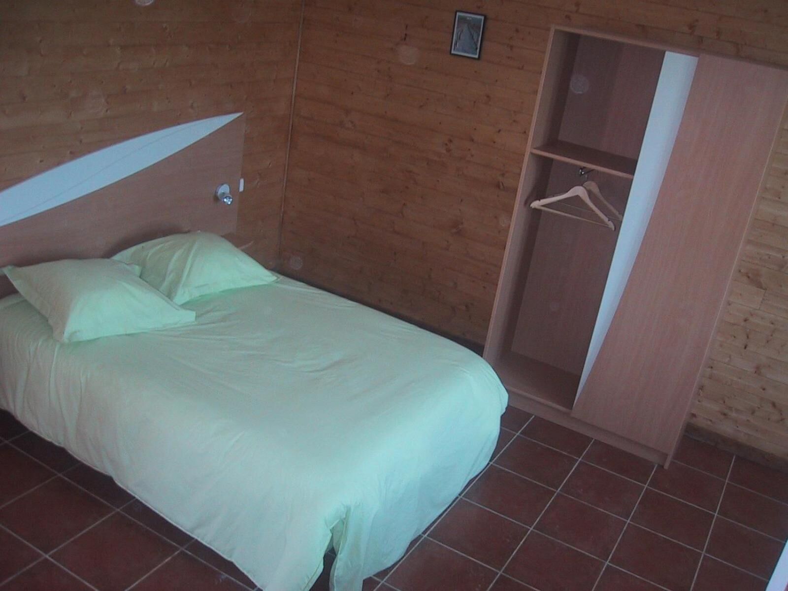 Un lit simple avec un matelas et des oreillers blancs, une armoire en bois et un sol carrelé.