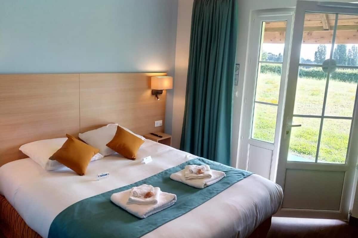 Ein Hotelzimmer mit Bett, Nachttisch, Lampe, einem Fenster mit Vorhängen und Blick auf ein grünes Feld.
