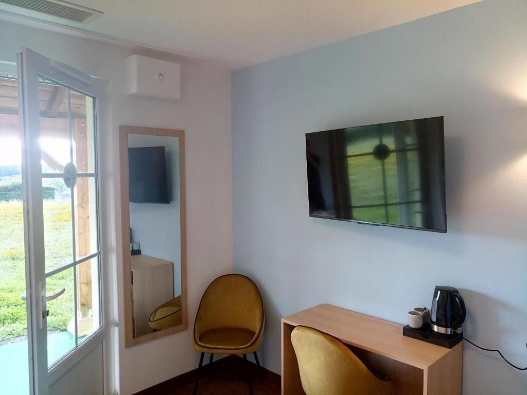 Ein Zimmer mit großem Fenster, einem an der Wand befestigten Fernseher, einem Holztisch und einem gelben Stuhl.