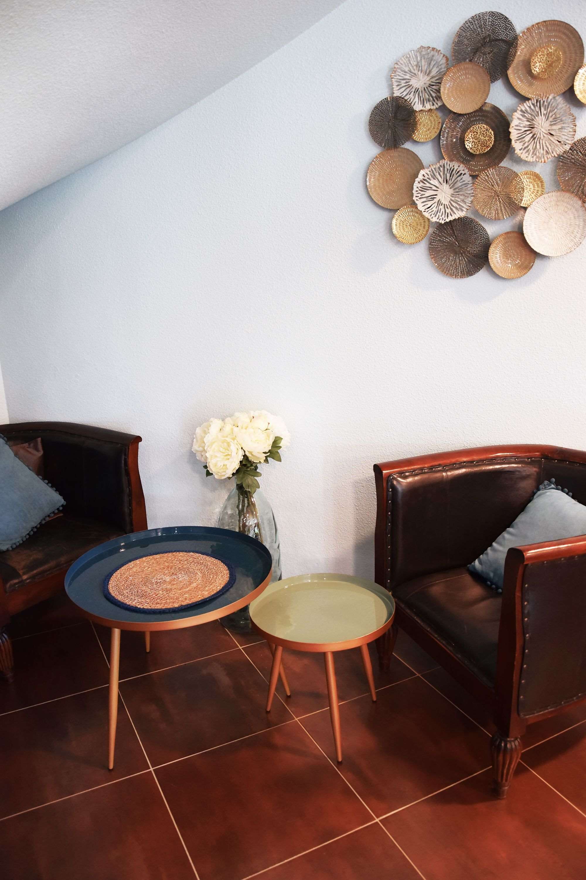 A room with two dark brown leather chairs, a small round table with a blue mat, and a vase with white flowers on a white wall.