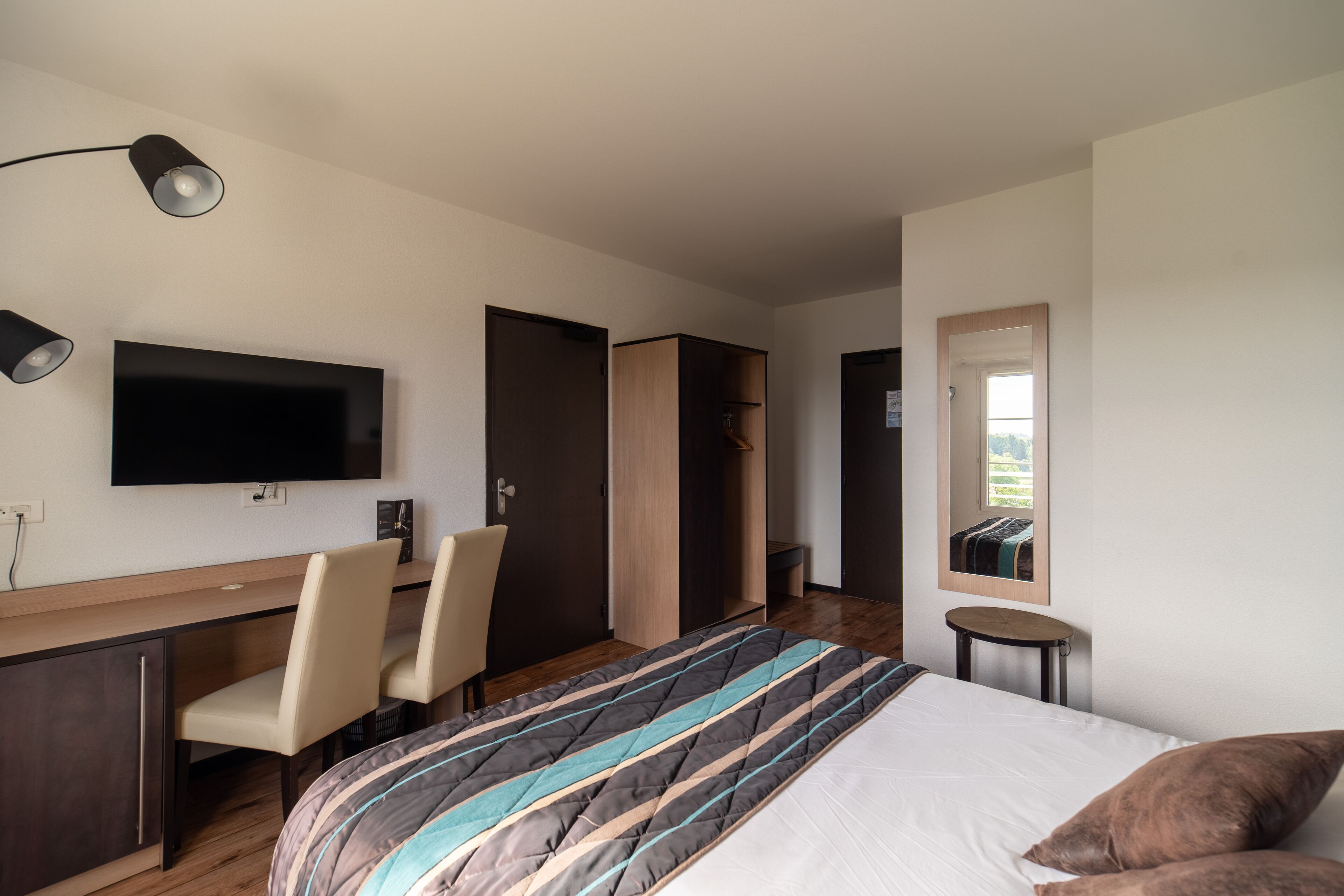 A modern hotel room with a bed, a desk with two chairs, a flat-screen TV, a wardrobe, and a small round table.