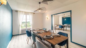 Een eethoek met een houten tafel voor zes personen, blauwe stoelen en uitzicht op het landschap door een raam met witte gordijnen.