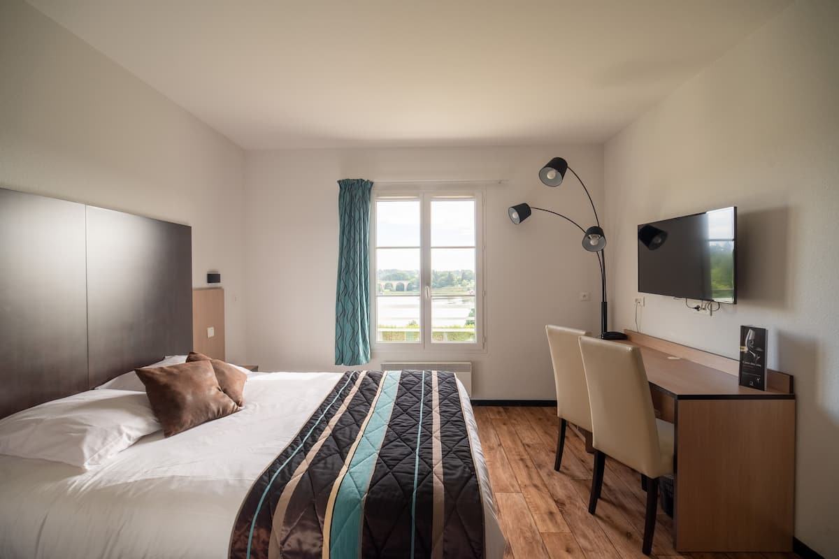 Ein modernes Hotelzimmer mit Bett, Schreibtisch, Stuhl, Fernseher und einem Fenster mit Blick auf Grünfläche.