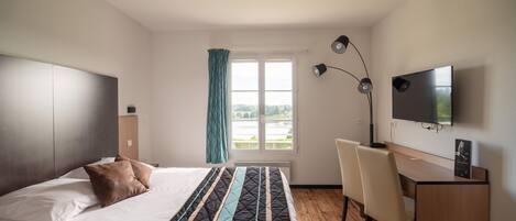 A modern hotel room with a bed, a desk, a chair, a TV, and a window with a view of greenery.