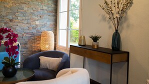 Een kamer met een stenen muur, een raam, een bank, een kleine tafel met een vaas, en een console met decoratieve objecten.