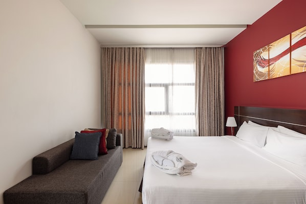A hotel room with a bed, a sofa, a window with curtains, and a wall with a painting. - Palmeiras Suite Hotel (Luanda)