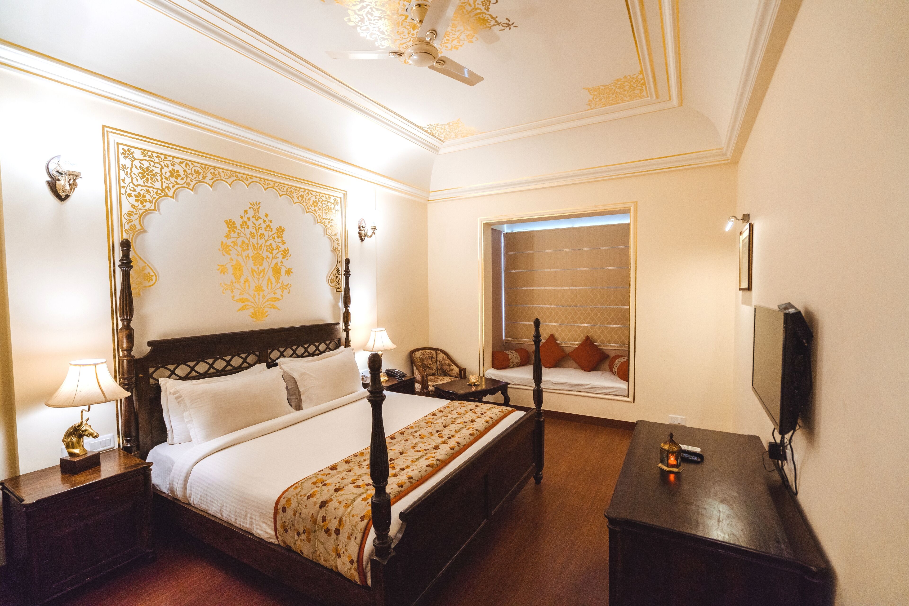 A hotel room with a four-poster bed, a desk with a lamp, a television, and a window with blinds.