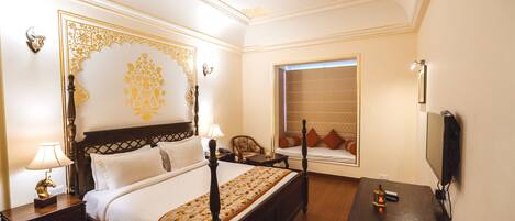 A hotel room with a four-poster bed, a desk with a lamp, a television, and a window with blinds.
