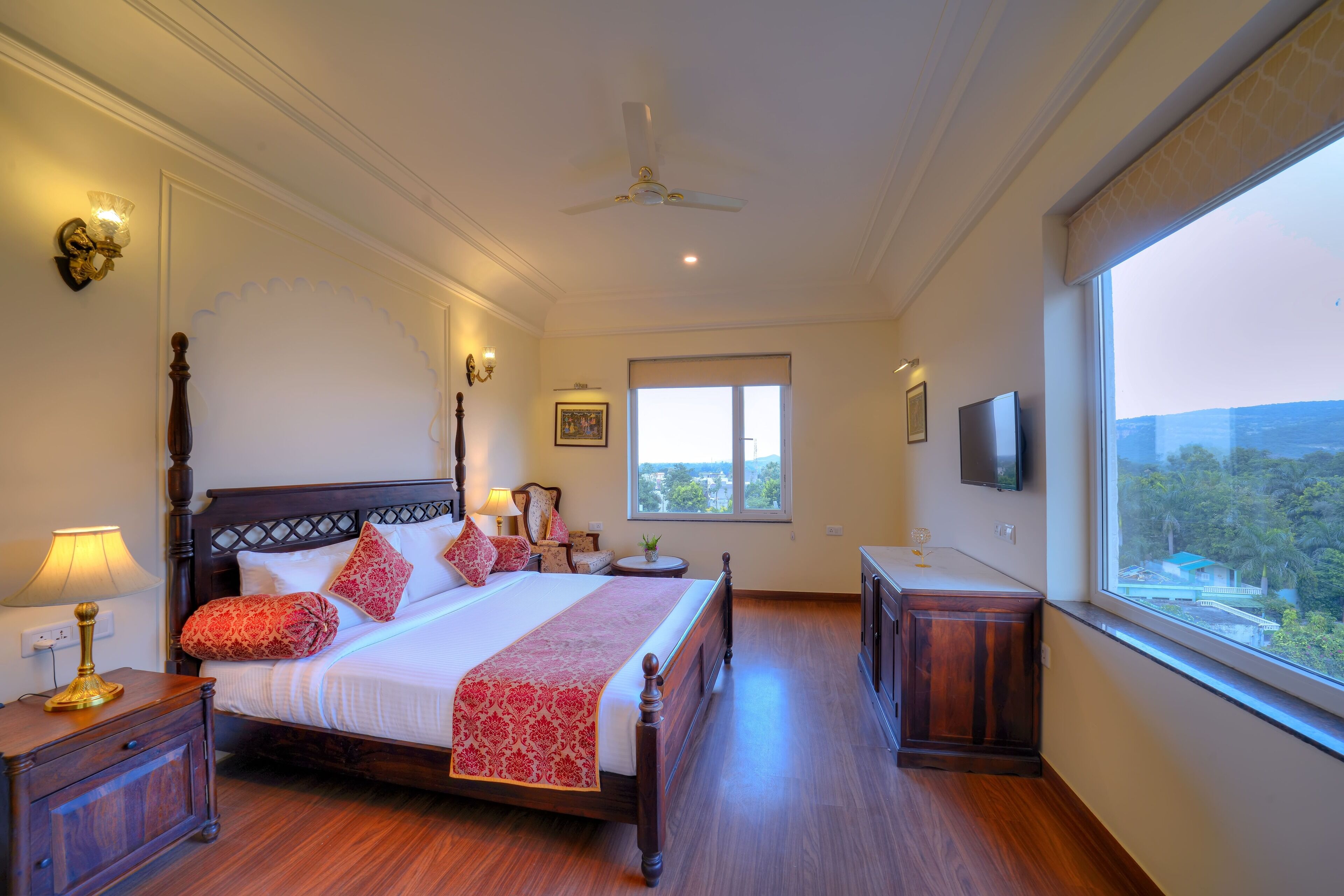 A bedroom with a large bed, a nightstand, a bedside table, a ceiling fan, a window with a view, and a TV mounted on the wall.