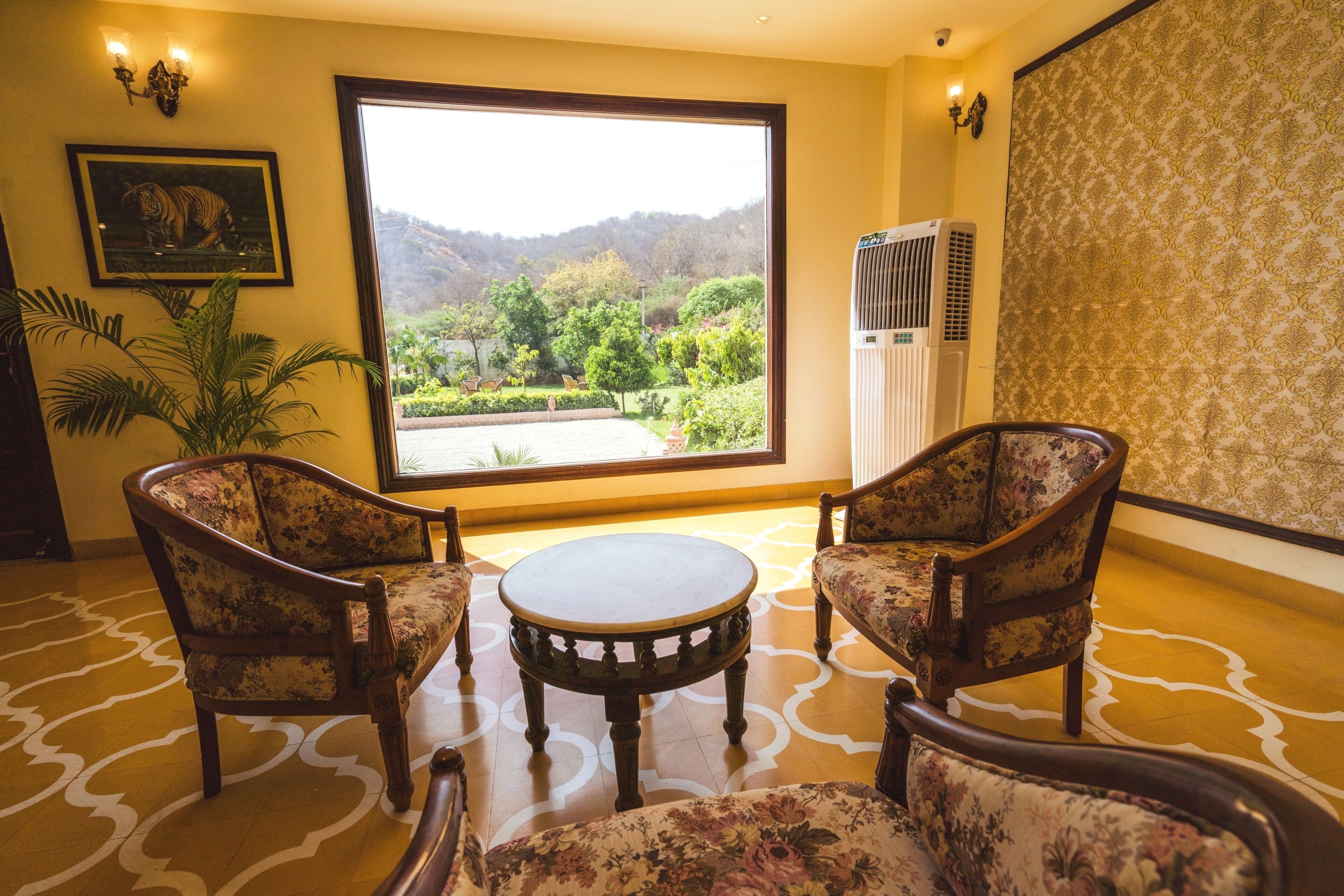 A room with two floral armchairs, a round wooden table, a large window with a view of greenery and hills, and a patterned carpet.