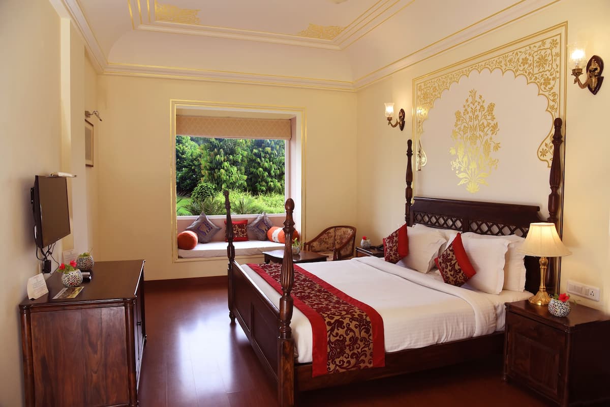 A bedroom with a large bed, a wooden nightstand, a desk with a lamp, and a window with a view of greenery.