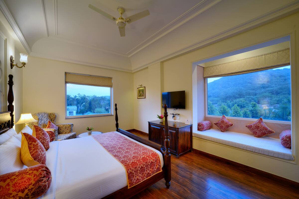 A hotel room with a large bed, a TV, a window seat with a view, and a ceiling fan.