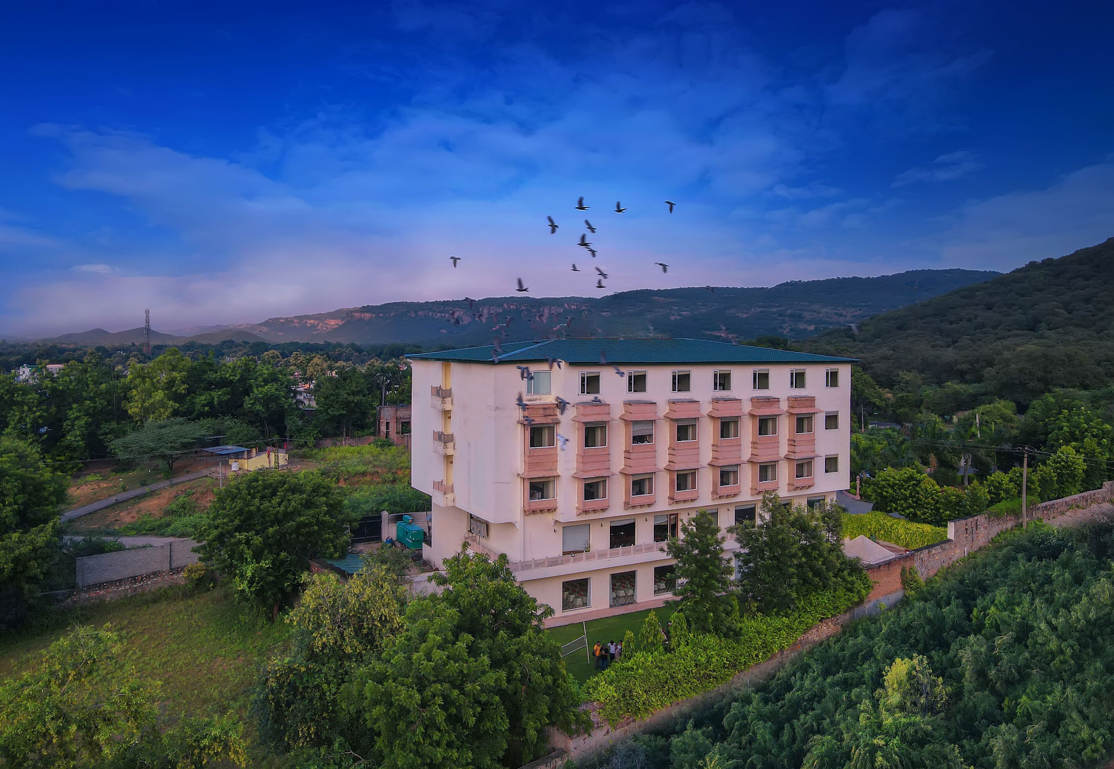 A multi-story building with a flat roof, surrounded by greenery and hills in the background.