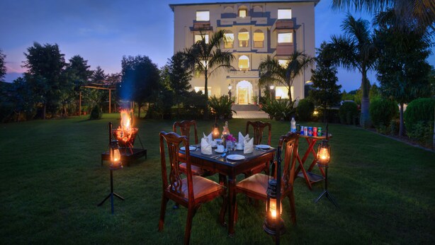 An outdoor dining setup with a table and chairs, surrounded by a well-maintained lawn and palm trees, with a large building in the background. - WelcomHeritage Mount Valley Resort, Ranthambore (Sawai Madhopur)