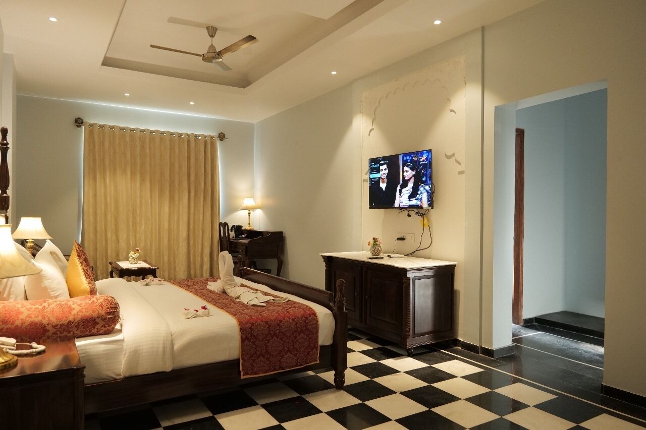A hotel room with a checkered floor, a bed with a red bedspread, a nightstand with a lamp, a TV mounted on the wall, and a ceiling fan.