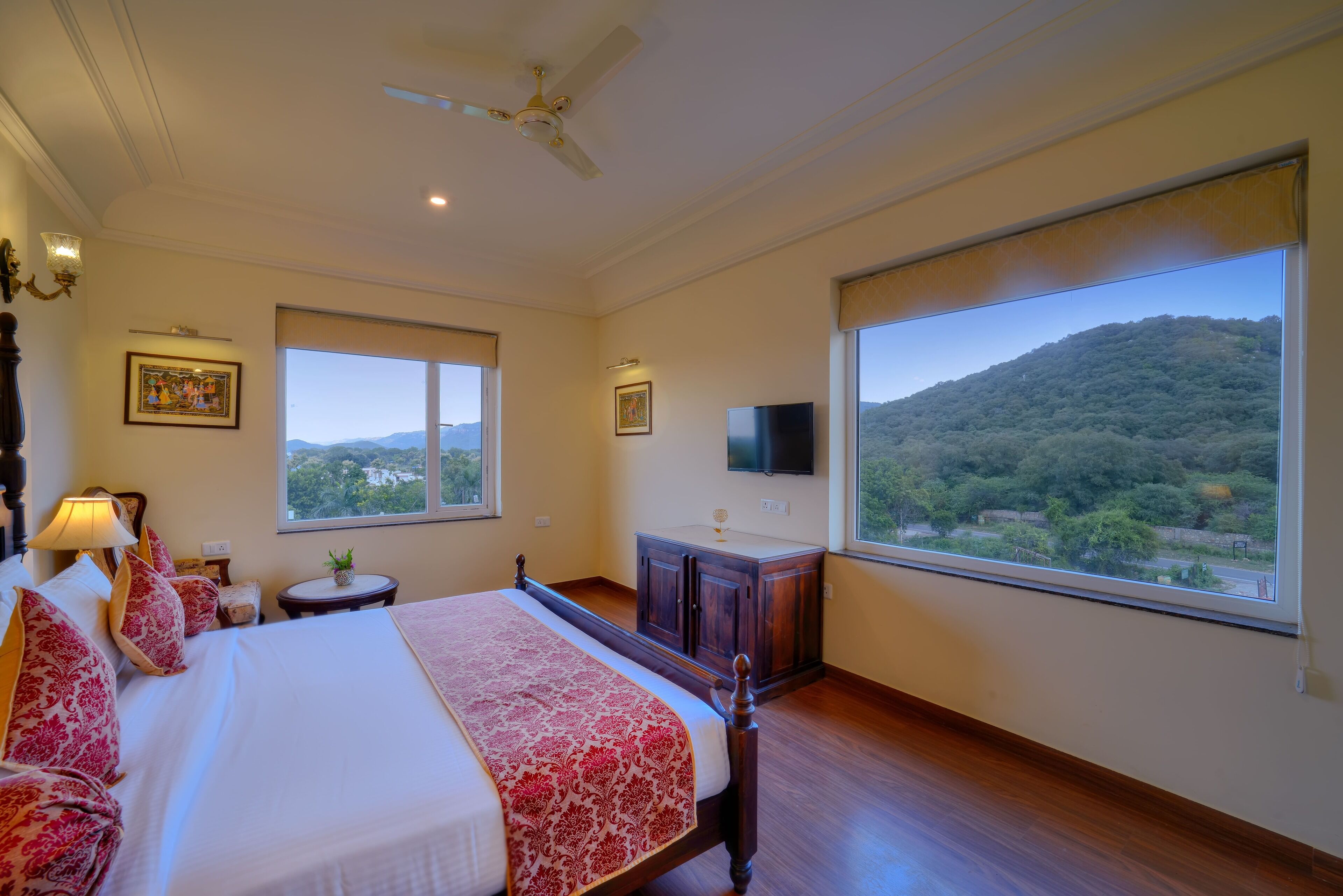 A spacious bedroom with a large bed, a ceiling fan, a TV, and a view of a mountainous landscape.