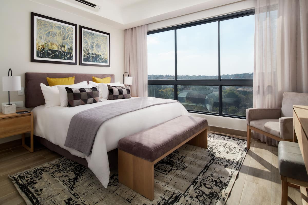 Ein modernes Hotelzimmer mit einem großen Bett, einer Bank, einem Sessel, einem Nachttisch und einem großen Fenster mit Blick auf die Stadt.