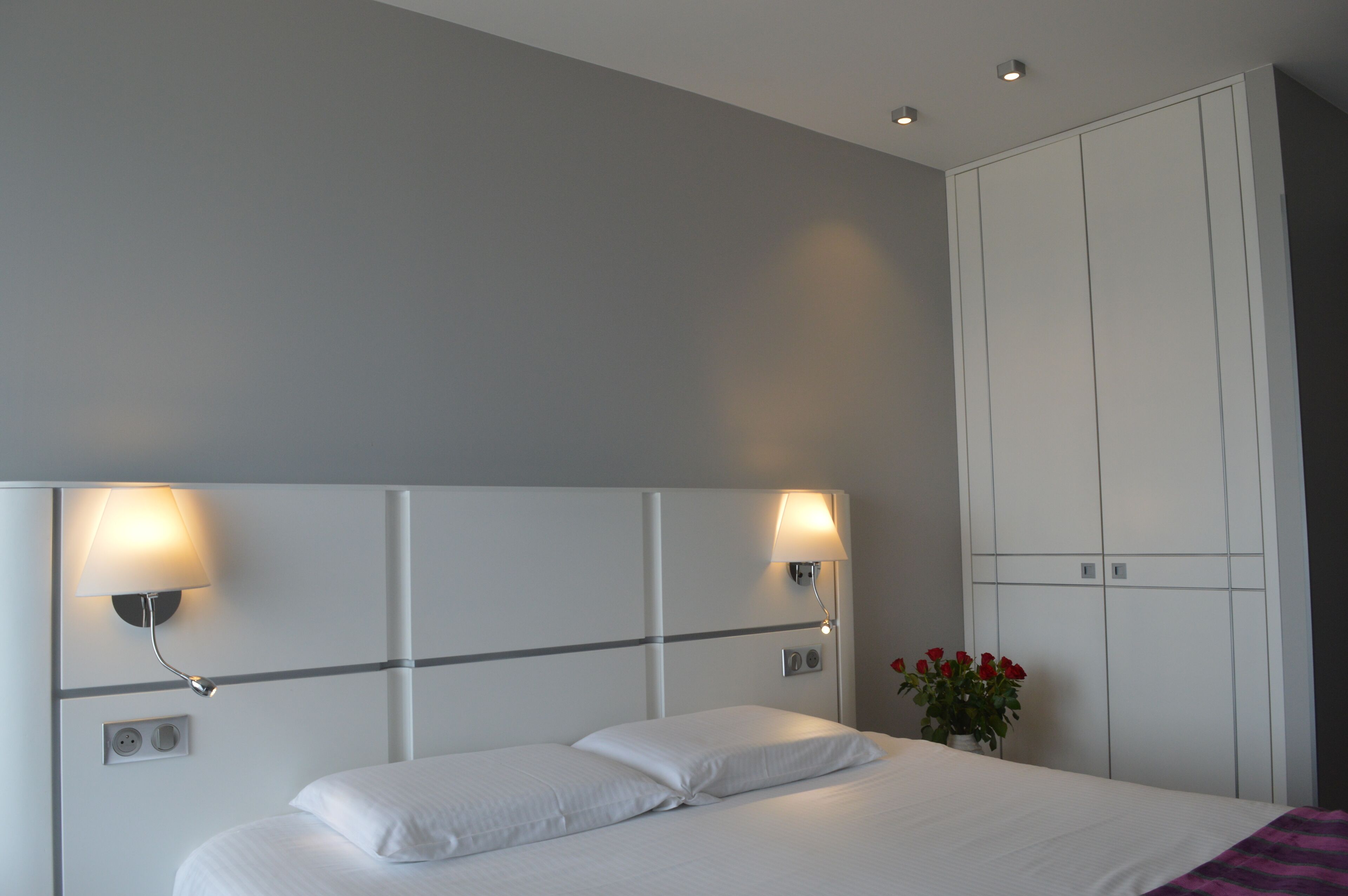 Habitación de hotel con una cama grande, dos lámparas de pared, un armario y una planta pequeña con flores rojas.