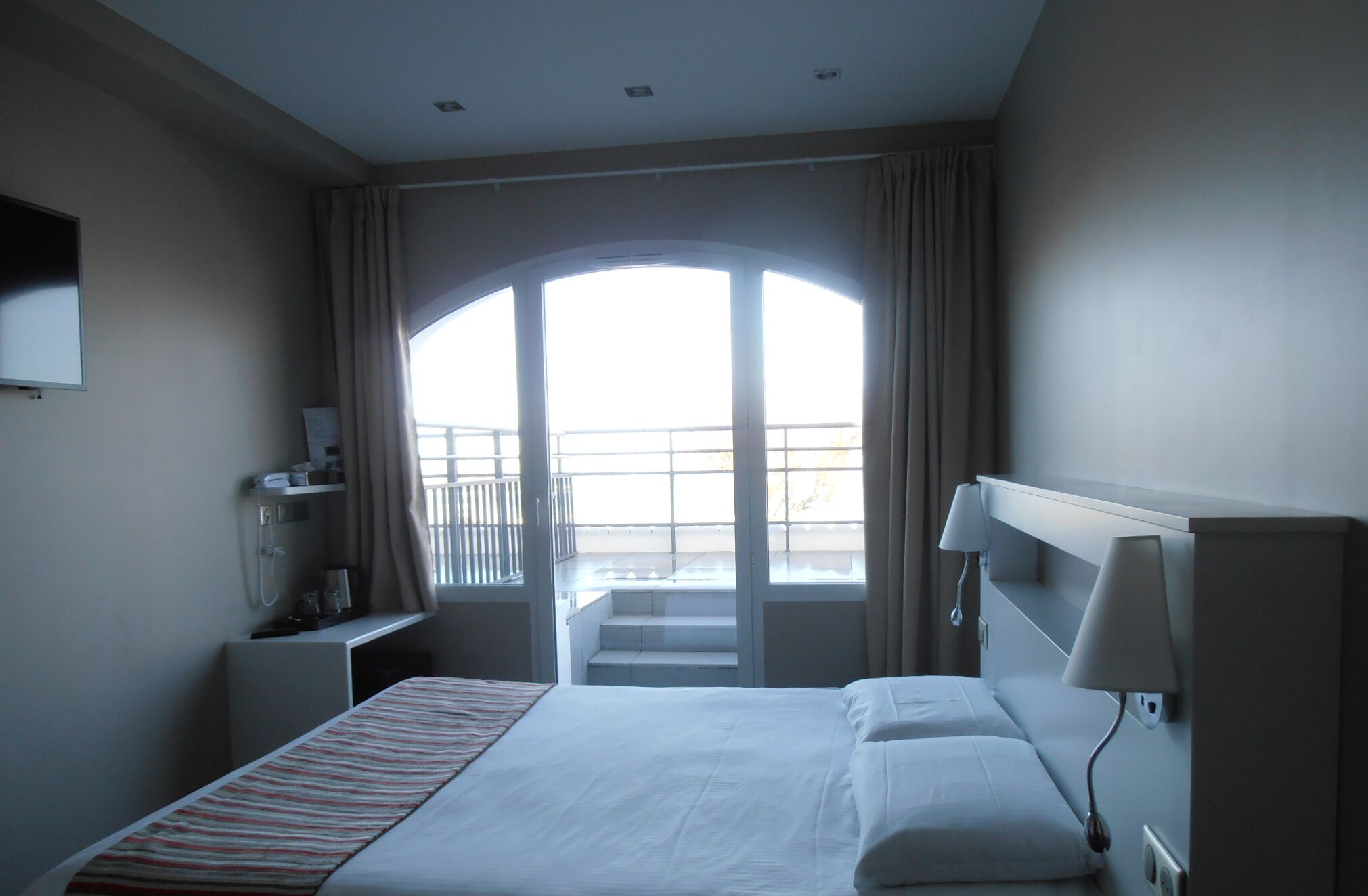 Habitación de hotel con una cama grande, un escritorio con lámpara, televisión en la pared y un balcón con vistas.