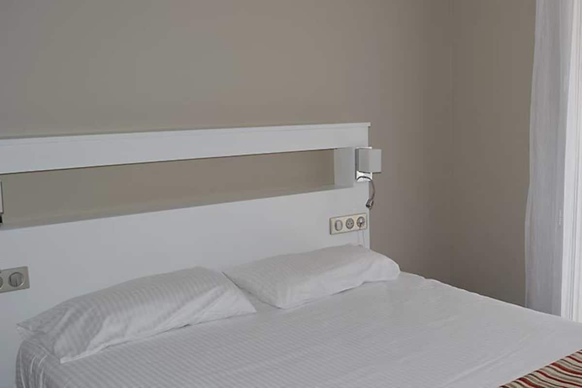 Una cama bien hecha con cabecera blanca, una mesita de noche con lámpara y una ventana con cortinas translúcidas.