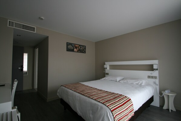 A bedroom with a bed, a bedside table, a wall-mounted light, and a door leading to another room.