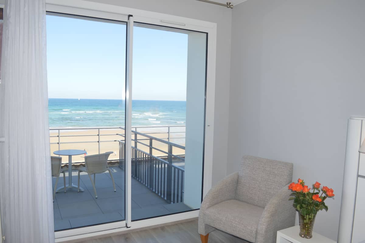 Habitación con puerta de vidrio corrediza que da a un balcón con vistas a la playa y al océano. Hay una silla y una mesita en el balcón.