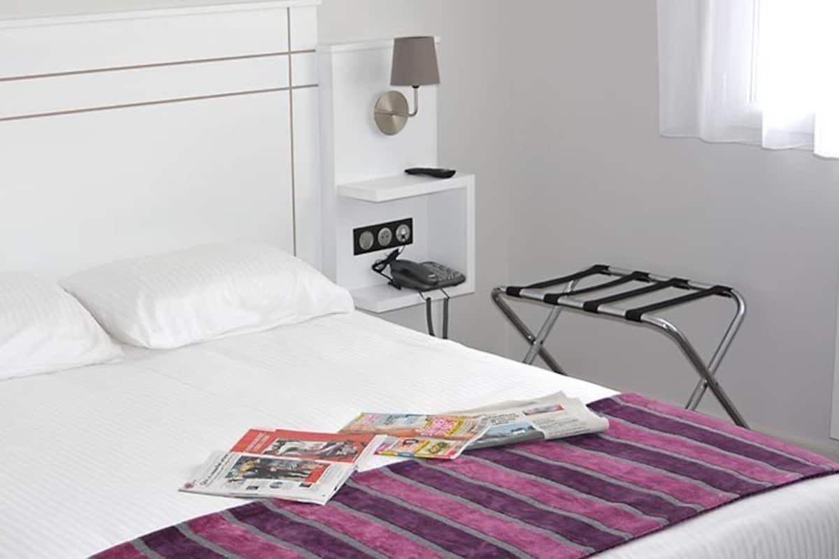 Una habitación de hotel con una cama, una revista sobre la cama, una lámpara en la mesita de noche y un perchero para toallas.