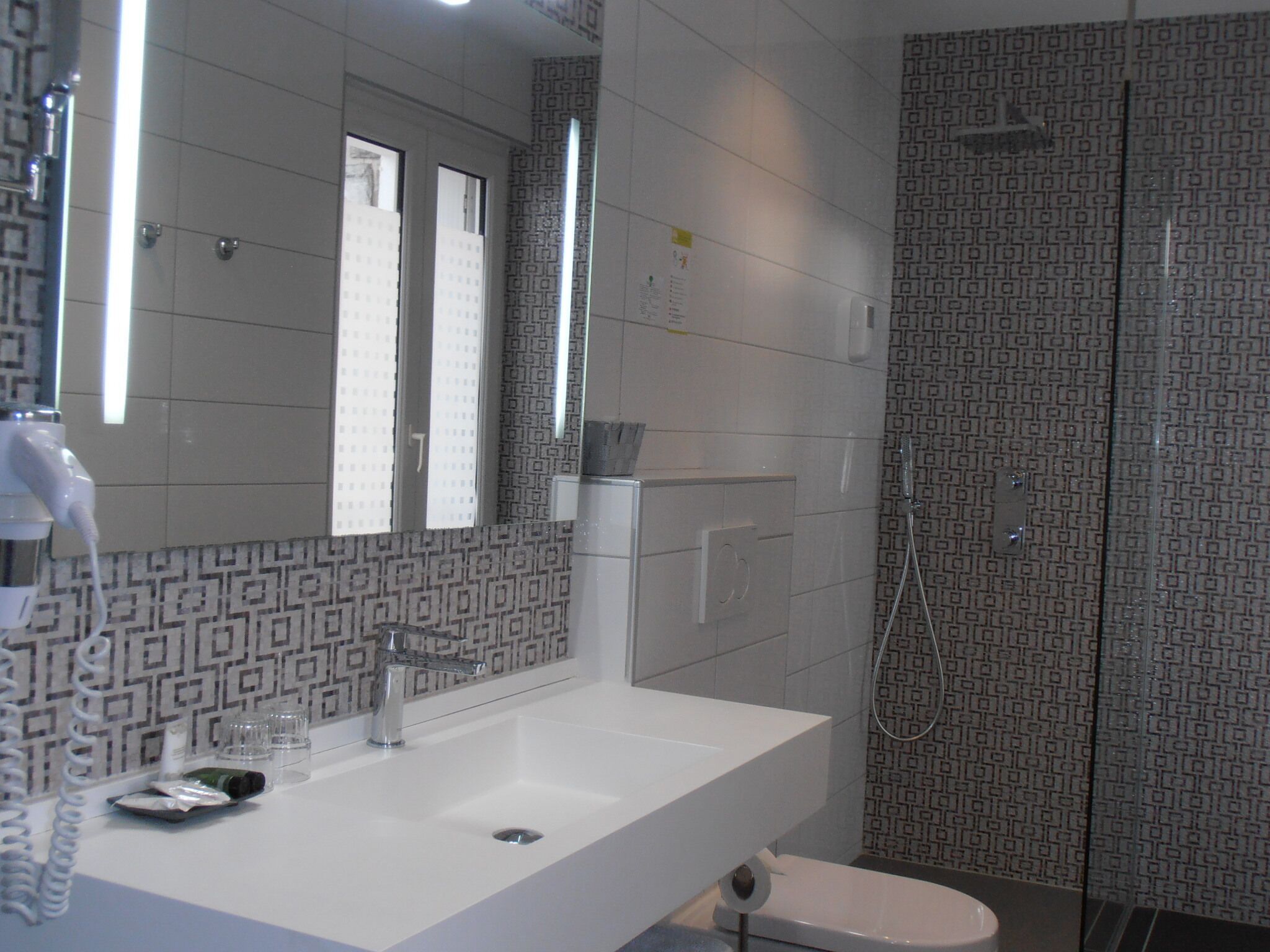 Un baño con lavamanos blanco, espejo, ducha y azulejos estampados.