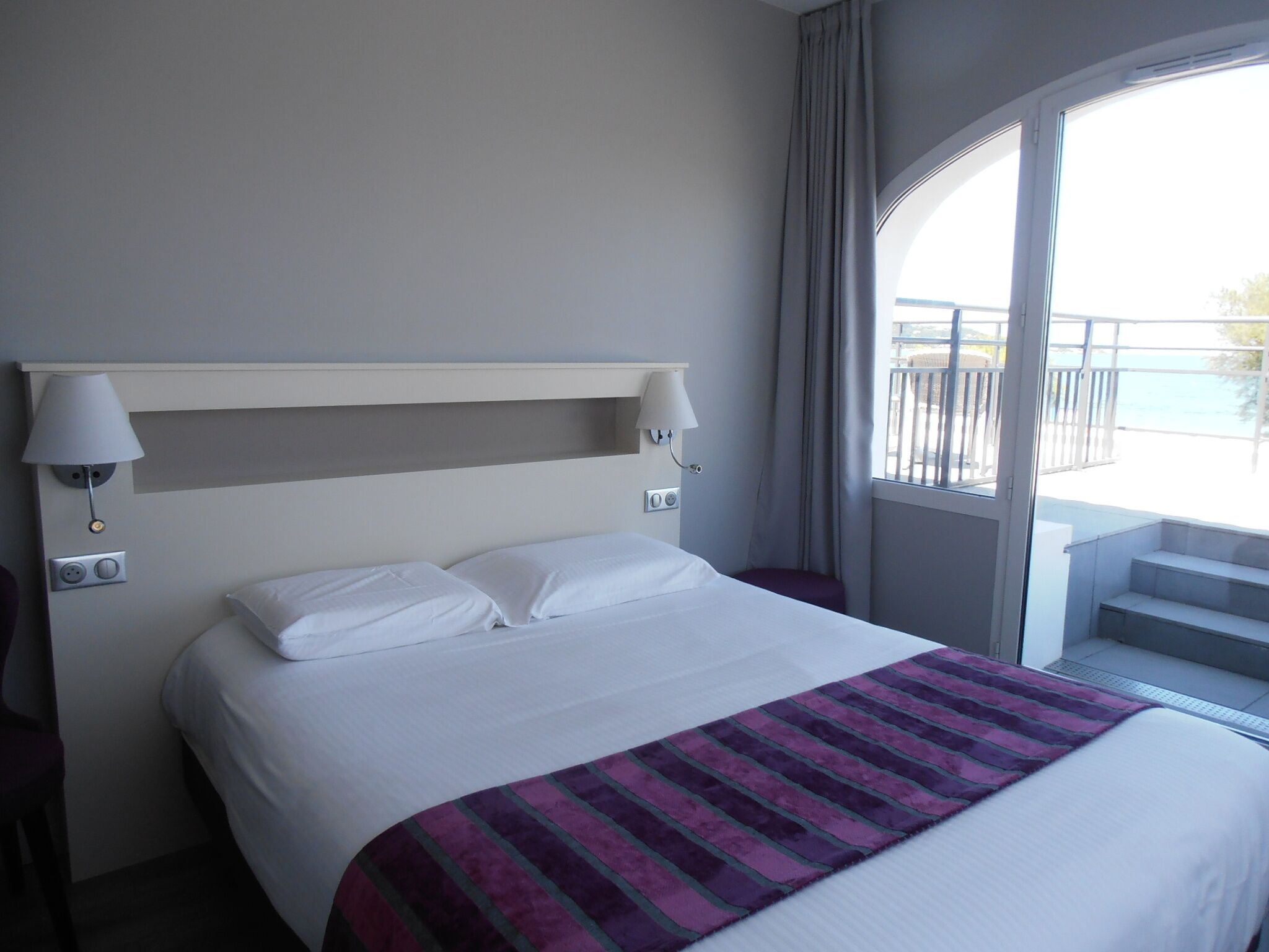 Habitación de hotel con cama, dos lámparas de noche, ventana con vistas y balcón.