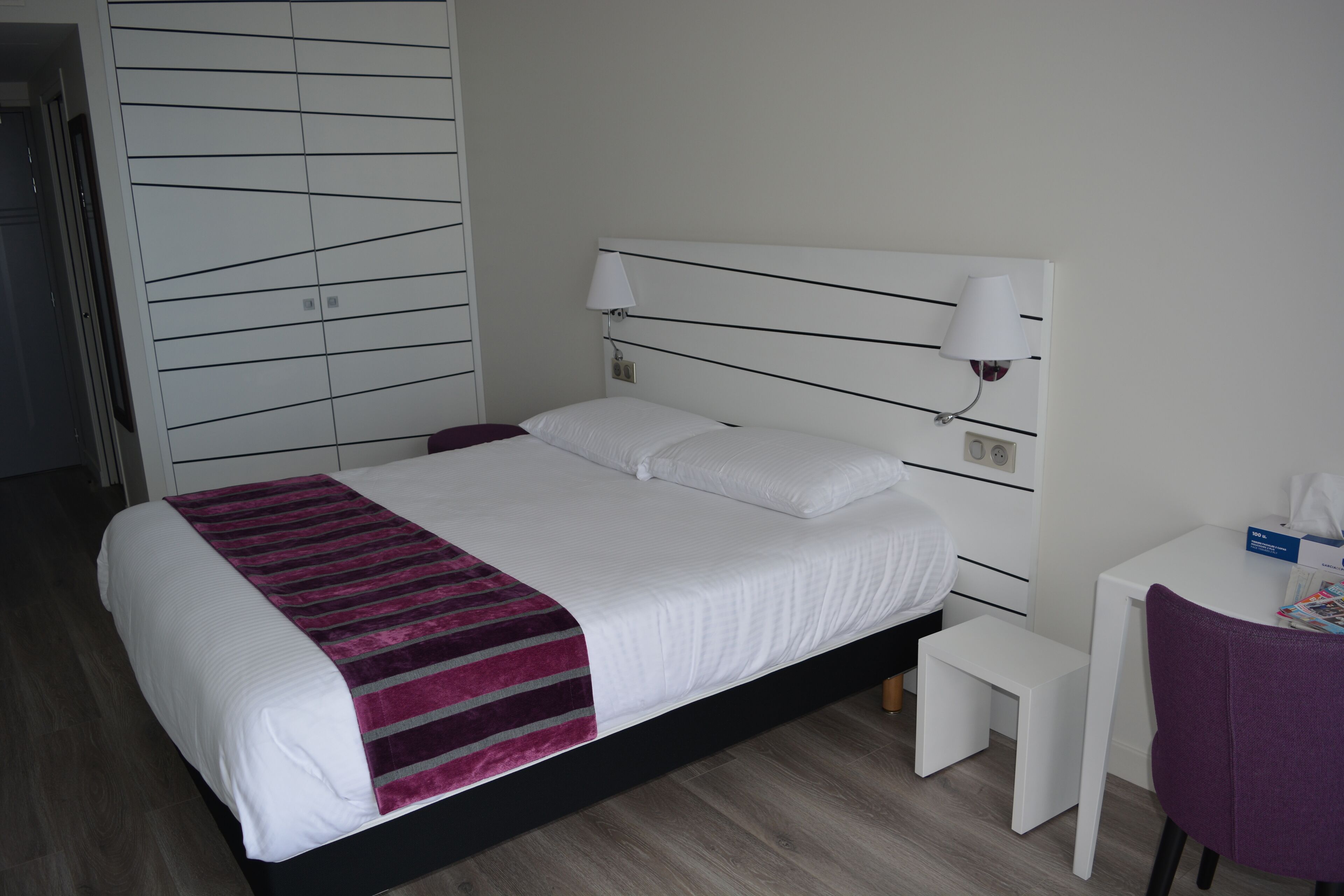 A neatly made bed with a white and purple striped blanket, a white bedside table with a lamp, and a small white stool.
