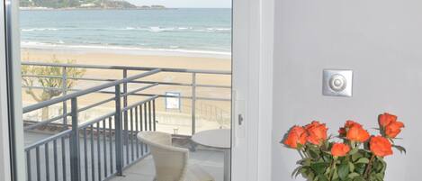 A balcony with a view of the beach, a chair, a table with a vase of flowers, and a door.