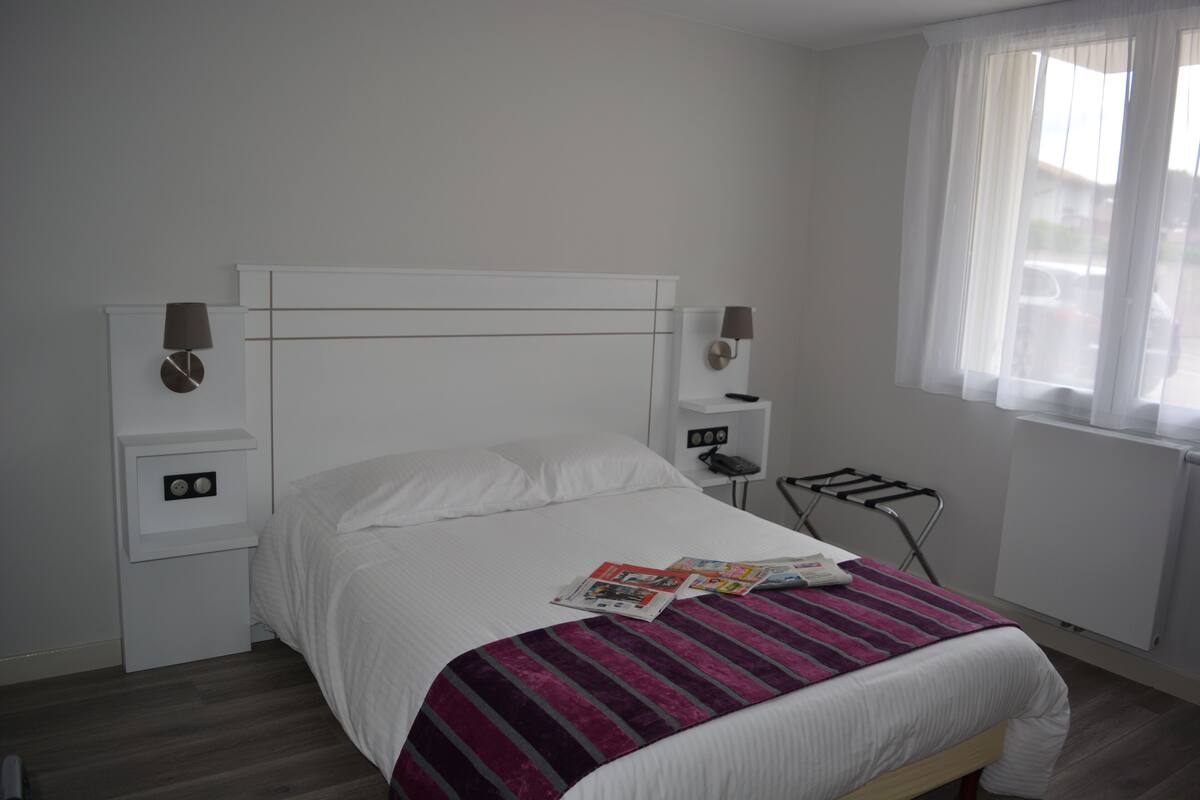 Una cama bien hecha con una manta a rayas, una mesita de noche con lámpara y una ventana con cortinas traslúcidas.