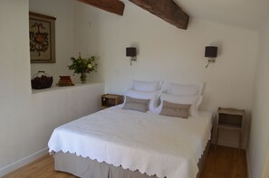 Un dormitorio con una cama grande, dos mesitas de noche, una lámpara de pared, un cuadro enmarcado y un pequeño estante con libros y una cesta.