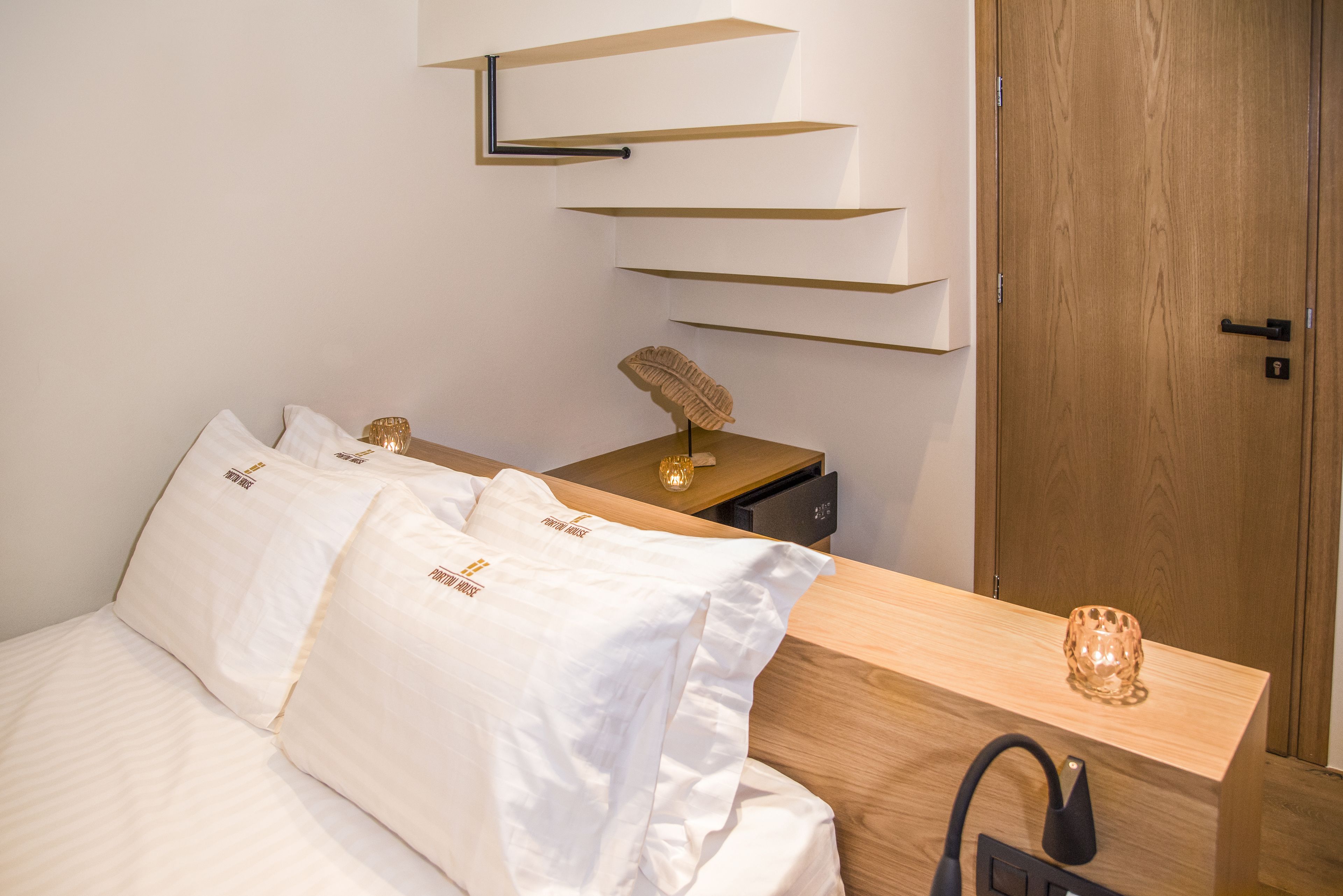 Un lit bien fait, avec du linge de lit blanc et des oreillers, une table de chevet en bois avec une lampe, et une étagère fixée au mur au-dessus du lit.