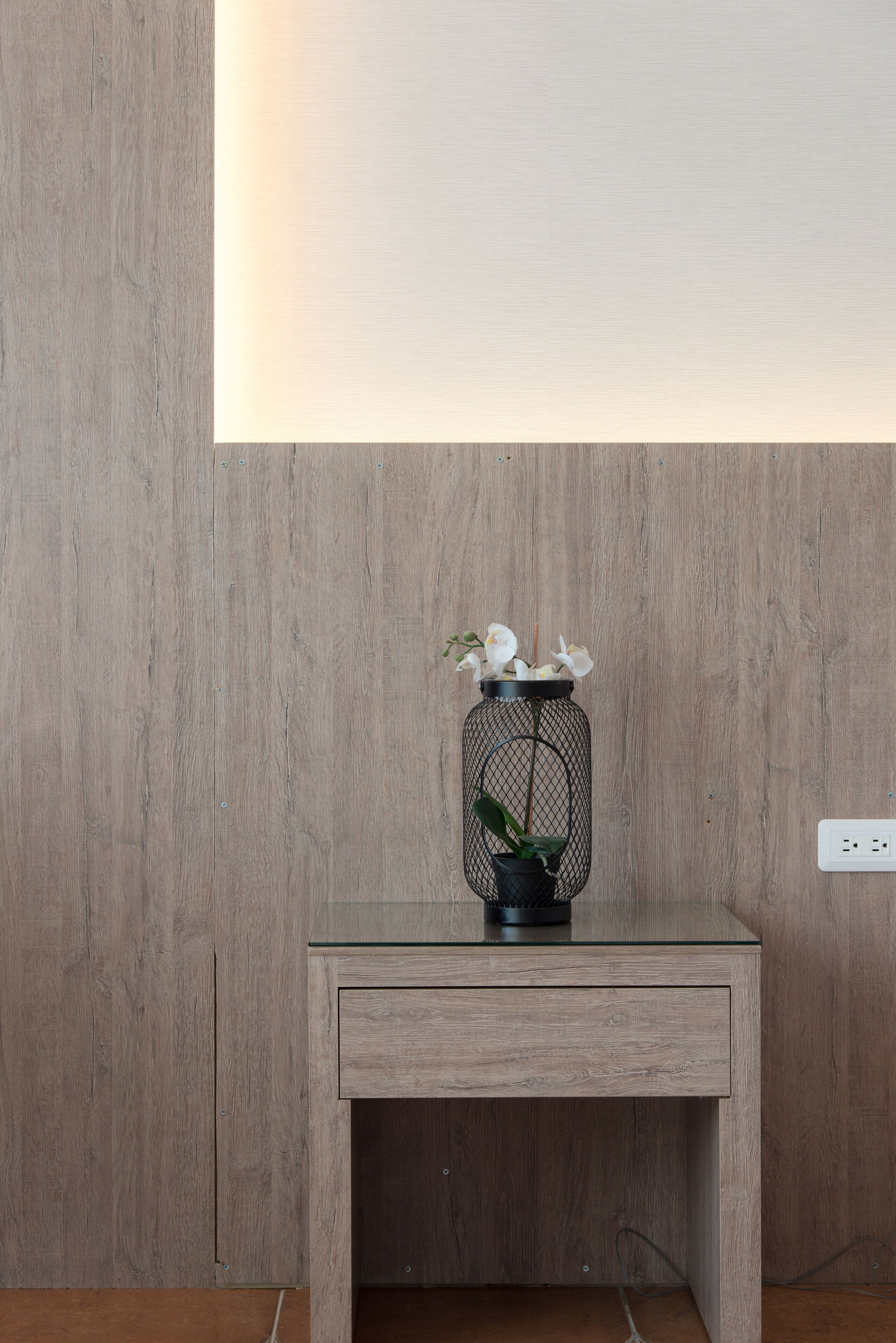 A wooden wall with a recessed light fixture, a small table with a glass top, and a wire basket containing white flowers.