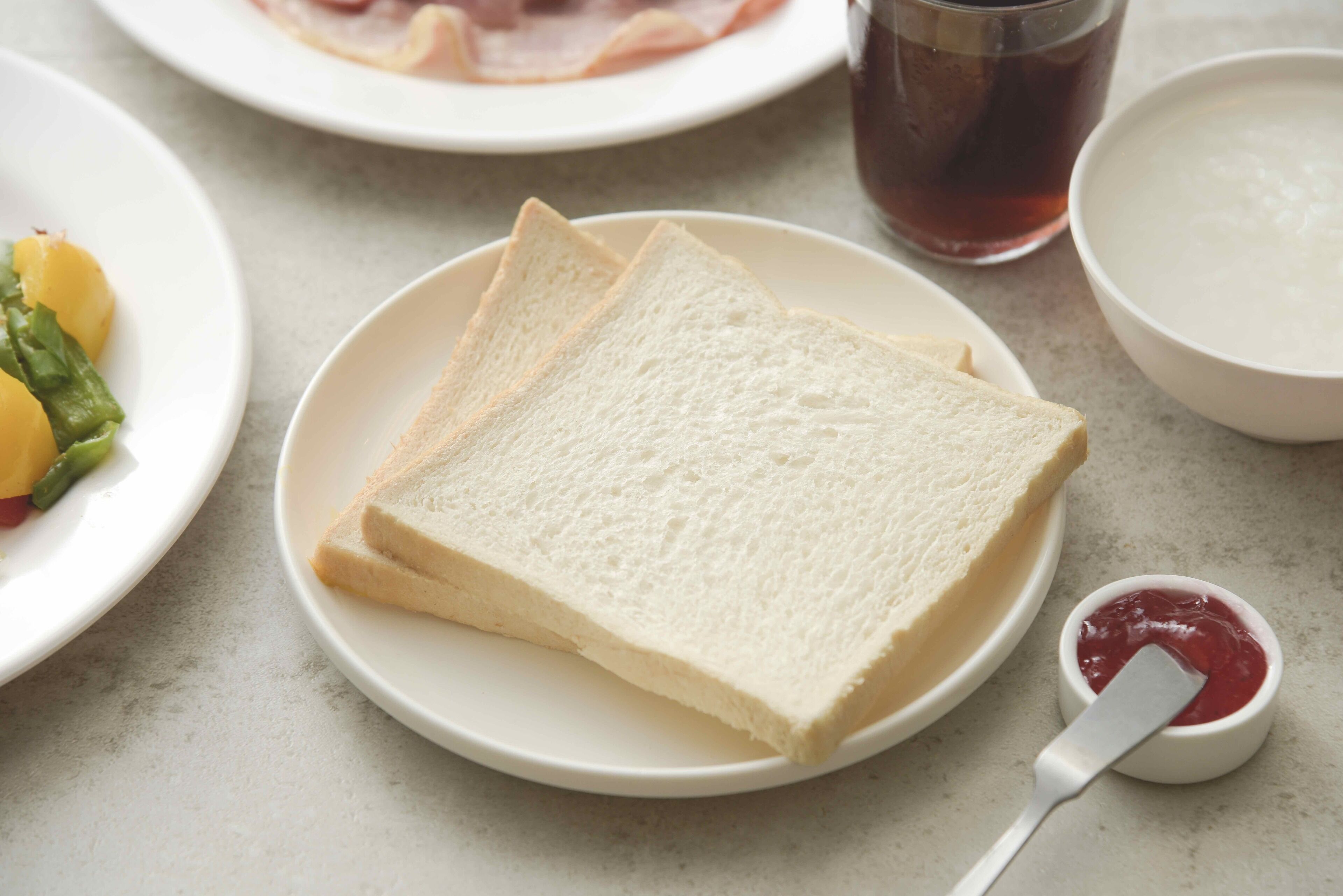 A plate with two slices of white bread, a small bowl of red jam, and a glass of dark liquid.