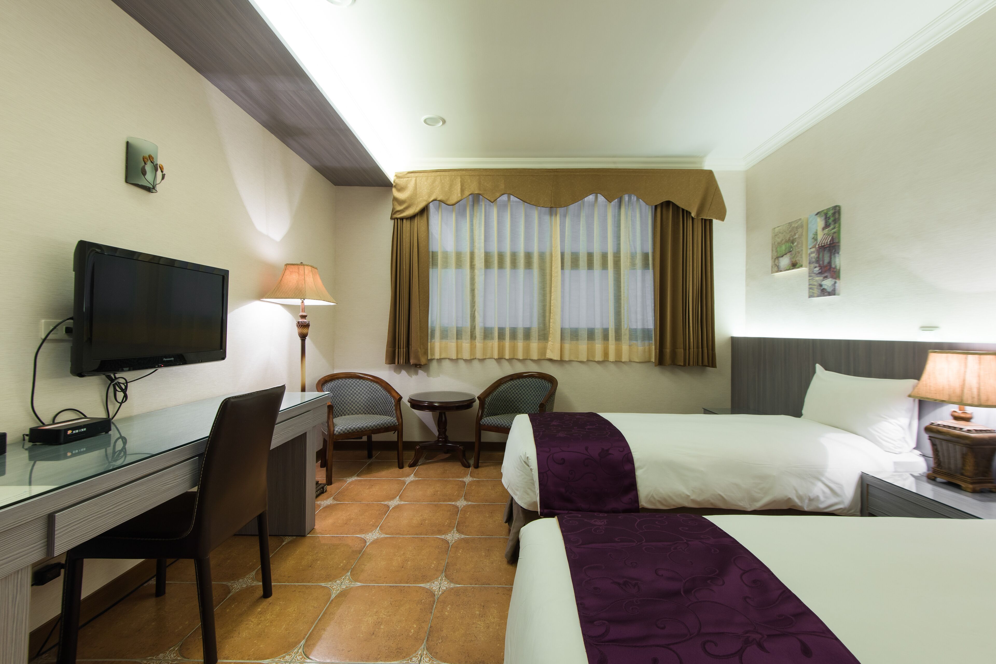 A hotel room with a bed, a desk with a television, two chairs, a small table, and a window with curtains.