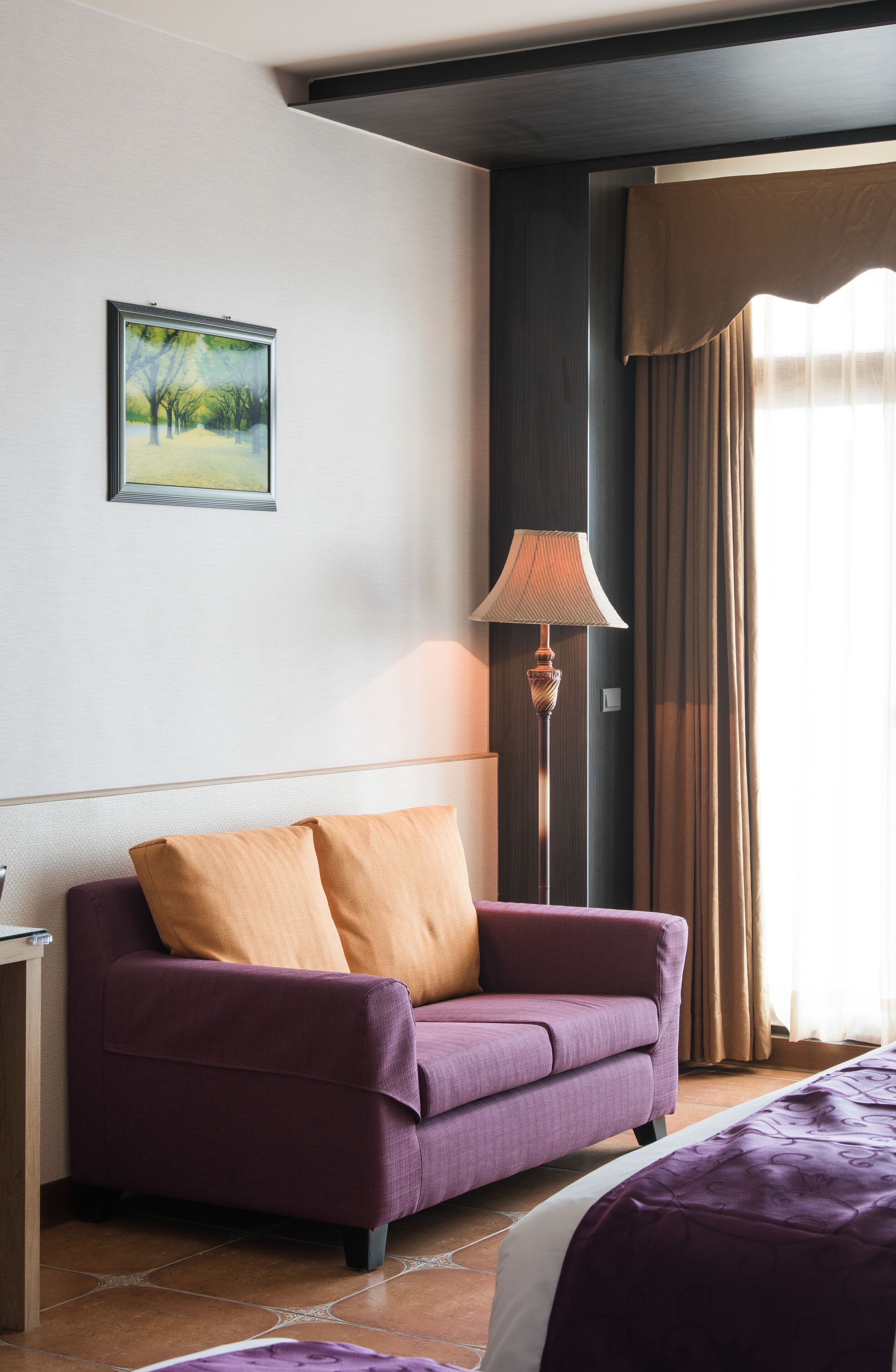 Een hotelkamer met een paarse bank, een bed, een vloerlamp en een schilderij aan de muur.