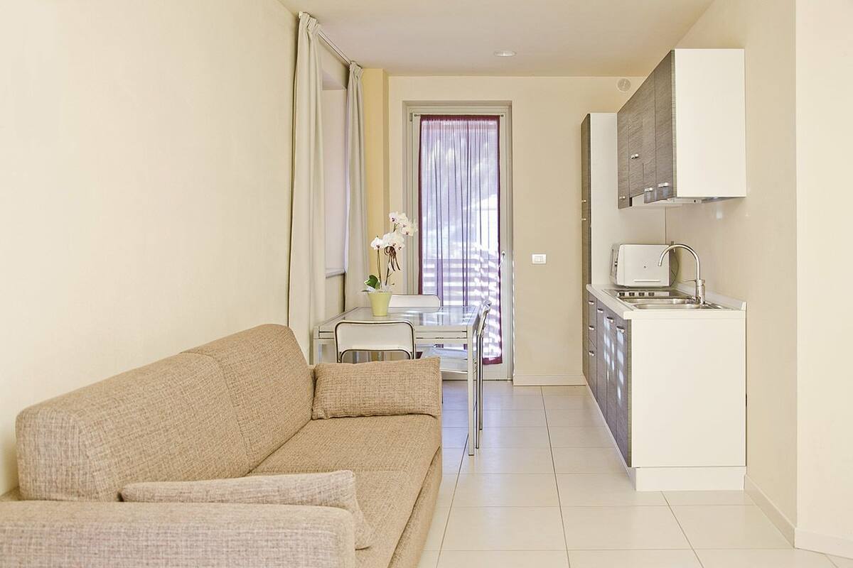 A compact living space with a sofa, a dining table with chairs, a small kitchen area with a sink and stove, and a window with curtains.