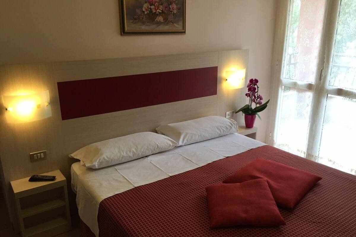 Habitación de hotel con cama, mesita de noche, lámpara de pared y ventana con cortinas.