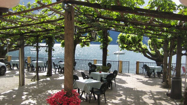 An outdoor seating area with tables and chairs, a pergola, and a view of the water. - Albergo Riva (Leggiuno)