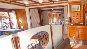 A hotel reception area with a wooden reception desk, a clock, and a view of the dining area. - Albergo Riva (Leggiuno)