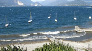 A coastal scene with sailboats on the water, a beach with pebbles, and mountains in the background. - Albergo Riva (Leggiuno)