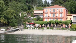A waterfront hotel with a red facade, multiple balconies, and a row of trees along the shore. - Albergo Riva (Leggiuno)
