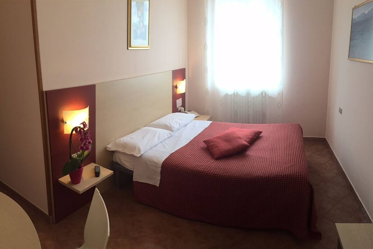 Habitación de hotel con cama, mesita de noche con lámpara, una silla y ventana con cortinas.