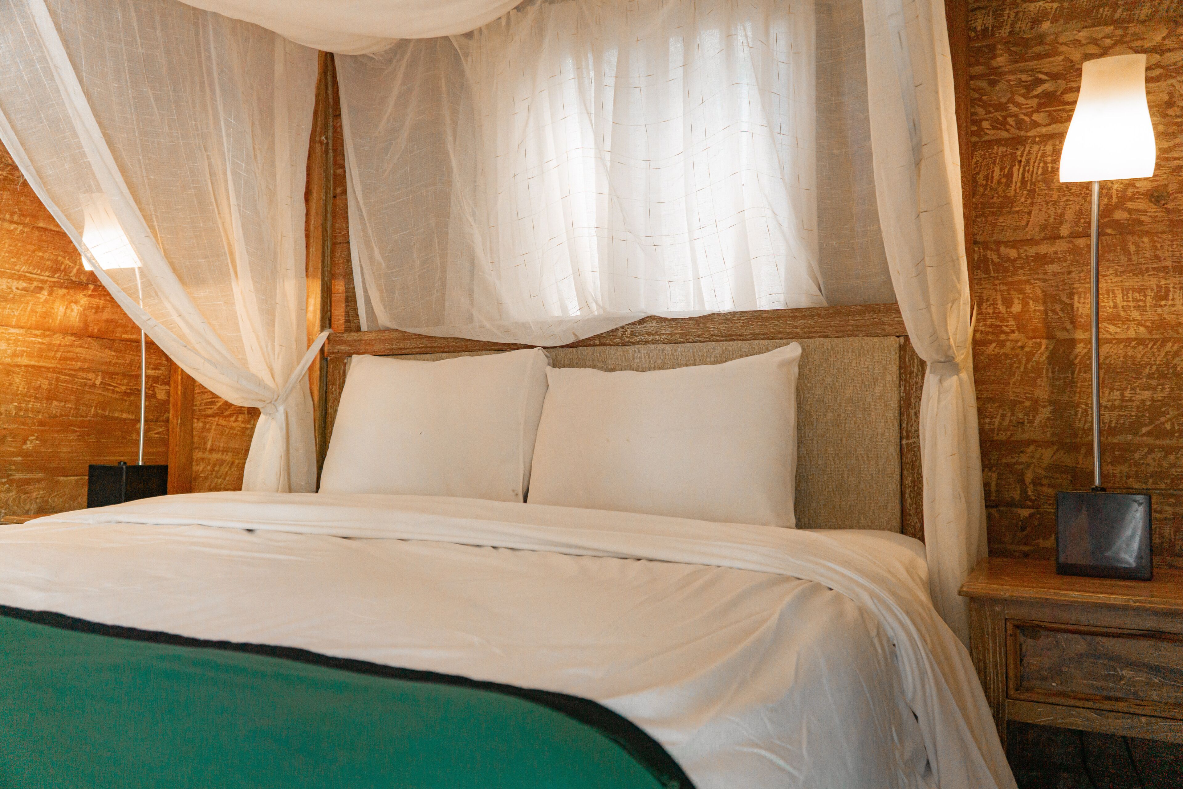 A bed with white linens and a green throw, a wooden headboard, and a nightstand with a lamp.