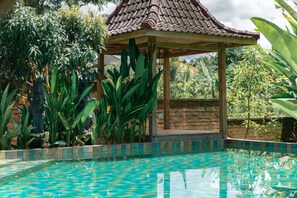 Ein Pool mit gefliestem Rand, umgeben von tropischen Pflanzen und einer Laube.